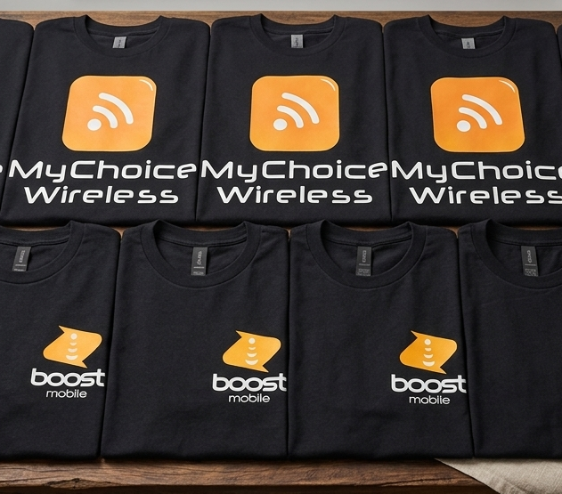 A neat row of black t-shirts featuring the orange "MyChoice Wireless" and "Boost Mobile" logos displayed on a wooden table with tech accessories like charging cables and a smartphone.
