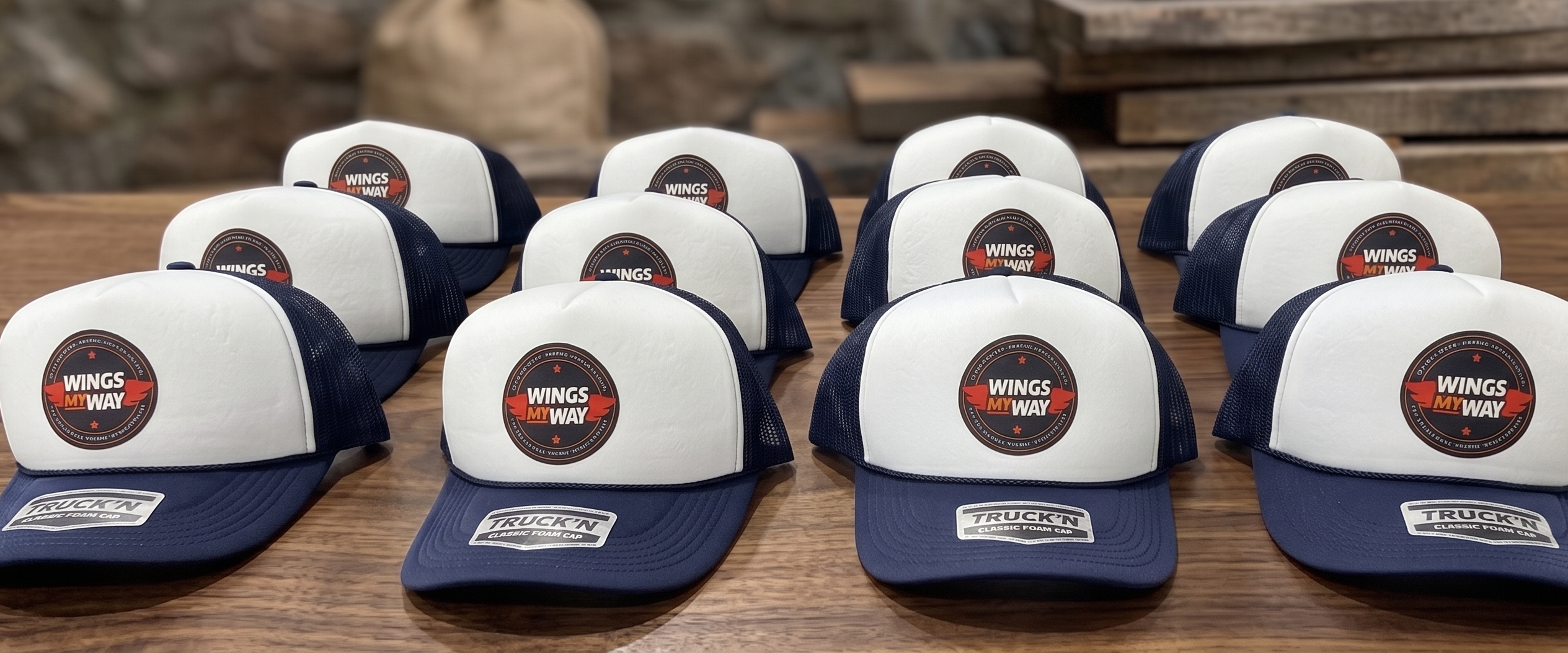 A collection of several navy blue and white with custom logo branded trucker hats arranged on a wooden surface against a rustic wood and stone background.