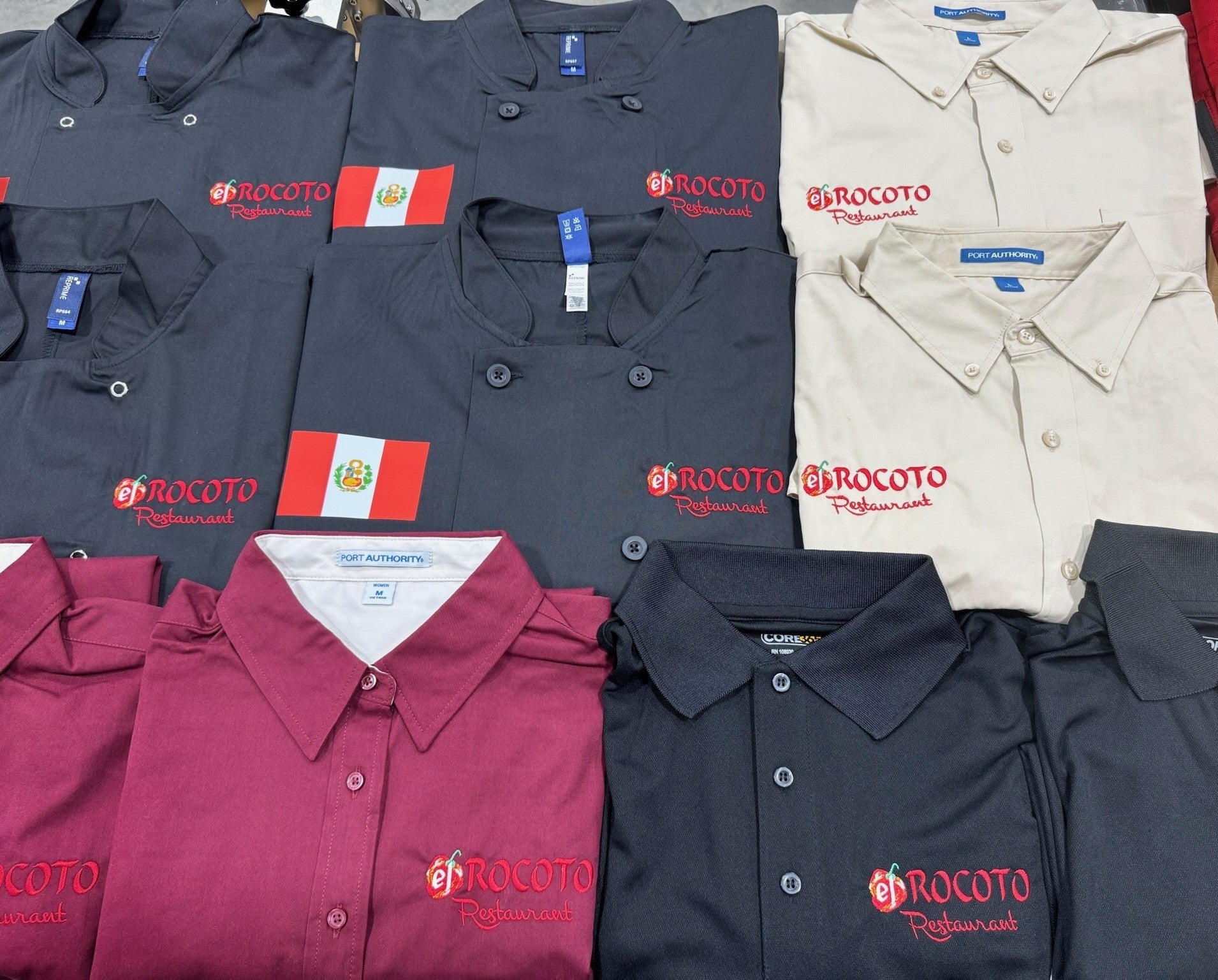 A neat collection of professional restaurant uniforms, including chef coats, polo shirts, and button-downs embroidered with a red restaurant logo on a warm tile background.